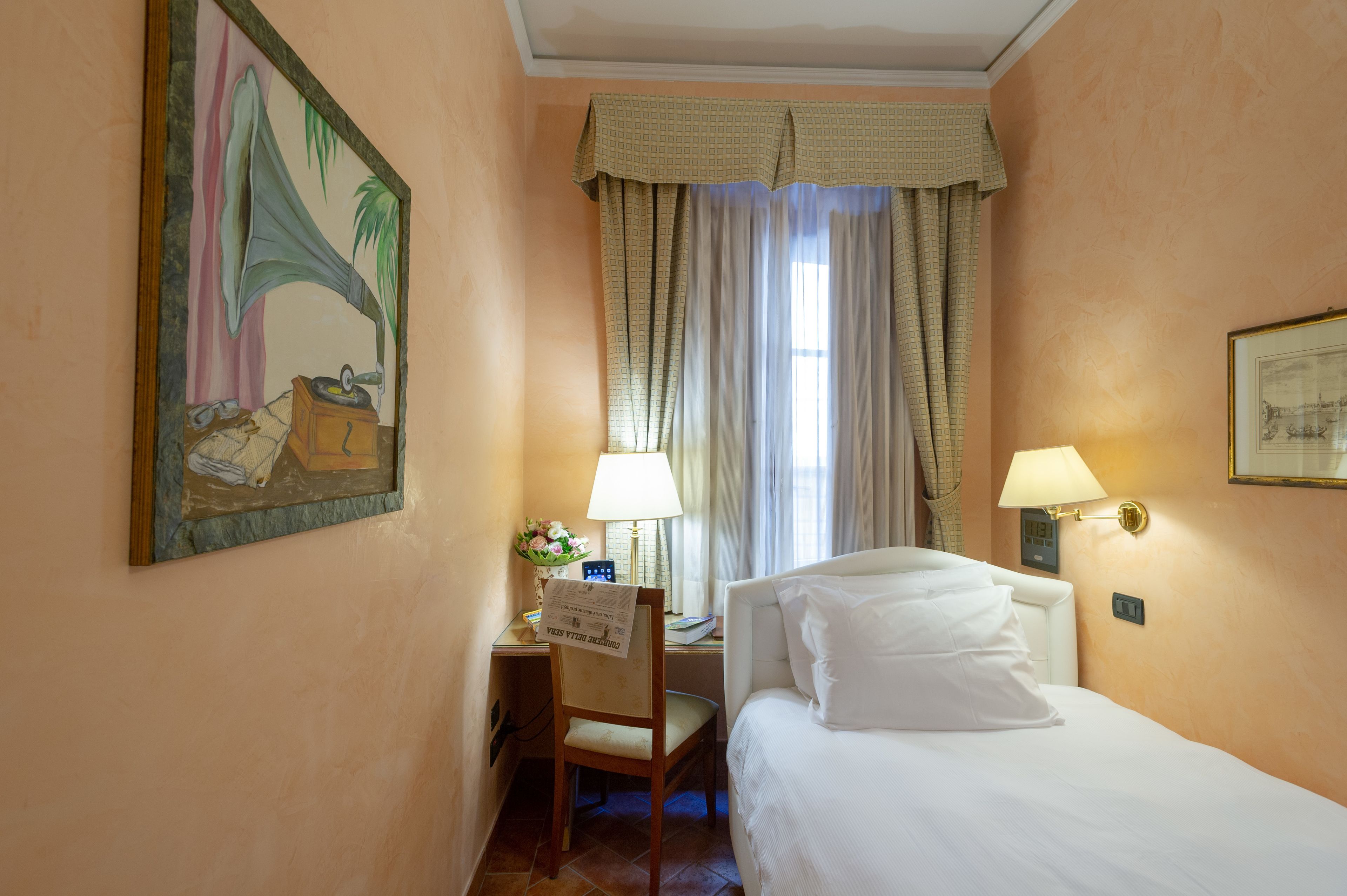 Habitación de hotel con cama, escritorio con lámpara, una silla, un cuadro en la pared y ventana con cortinas.