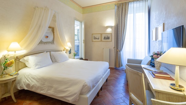 A hotel room with a large bed, a desk with a chair, a lamp, and a window with curtains. - Davanzati (Florence)