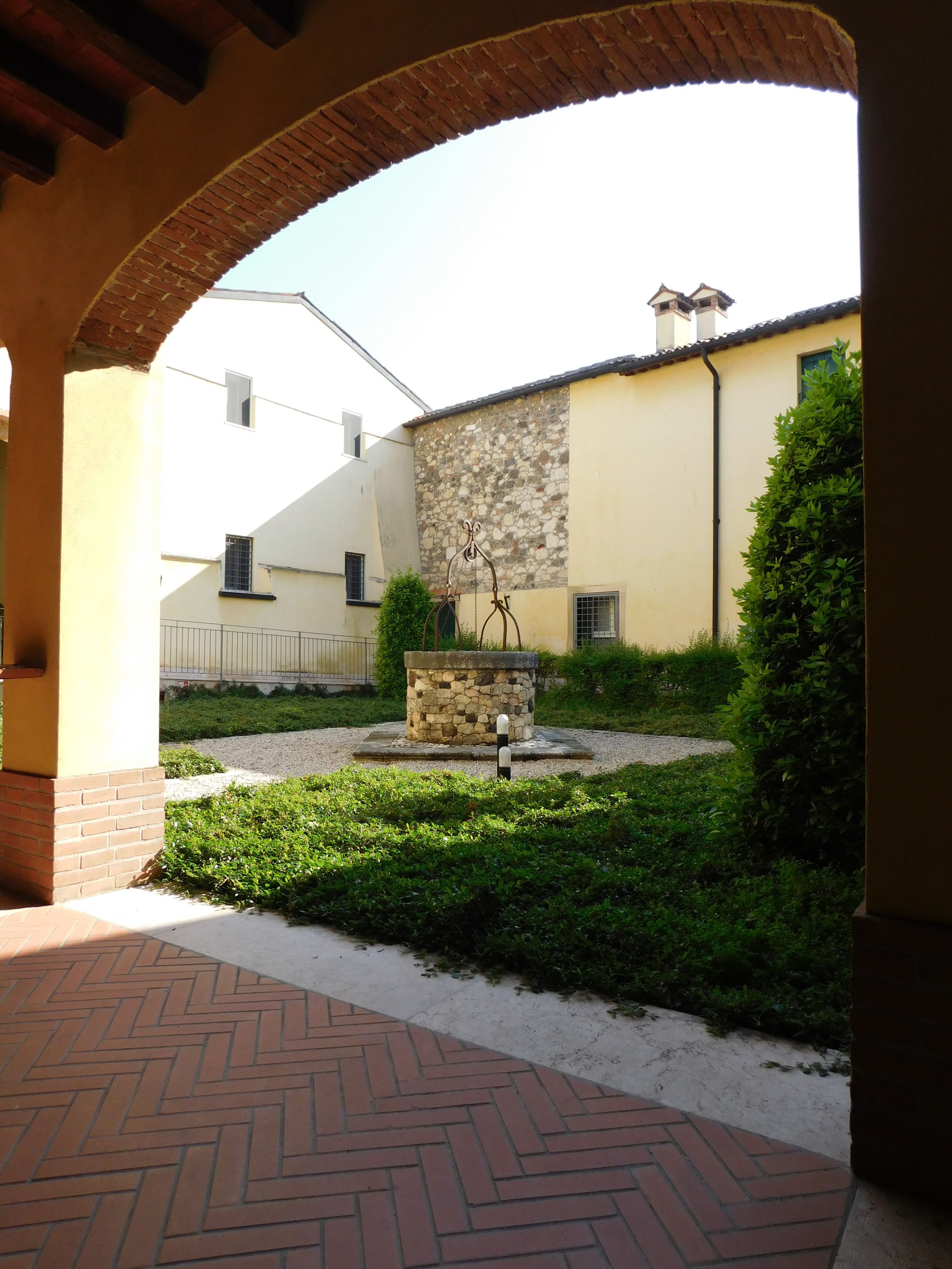 An outdoor courtyard with a stone fountain, a brick pathway, and a building with a stone wall and a tiled roof.