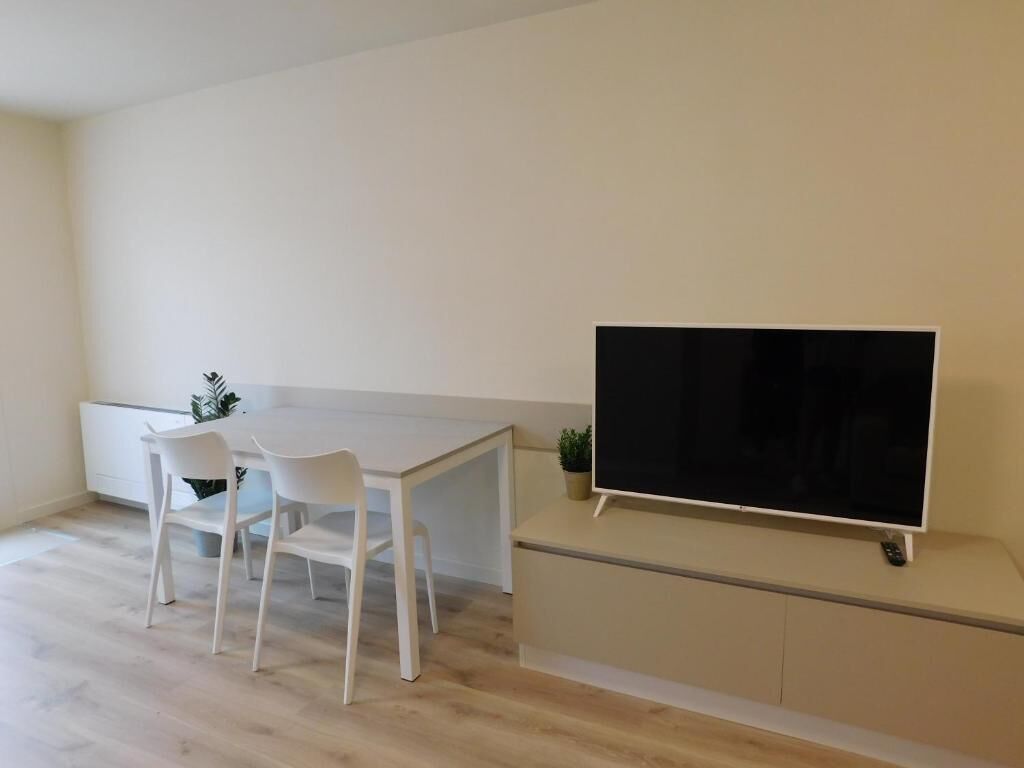 A modern living room with a flat-screen TV mounted on a cabinet, a white dining table with chairs, and a small potted plant.
