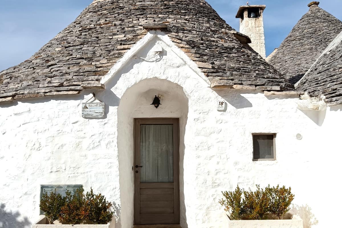A white building with a stone roof, a wooden door, and two potted plants.