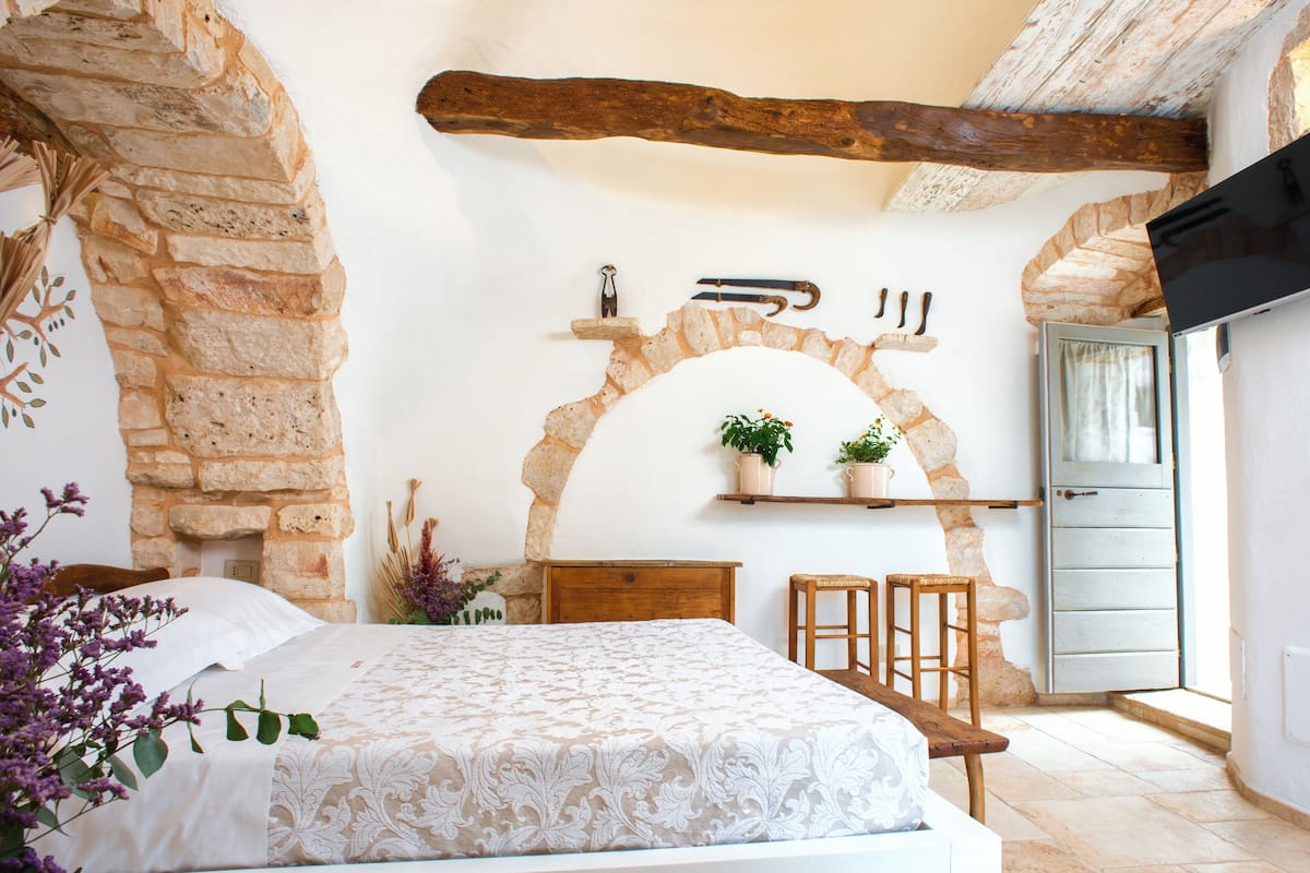 A bedroom with a stone fireplace, wooden beams, a bed with a patterned bedspread, a wooden nightstand, and a small table with chairs.