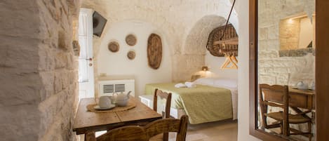 A cozy room with a stone wall, a wooden table with a teapot, a bed with a green bedspread, and a rustic wooden chair.