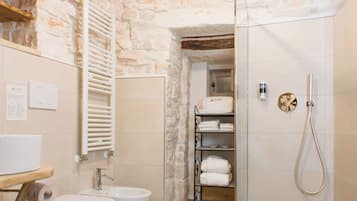 A bathroom with a stone wall, two toilets, a sink, a shower, and a towel rack.