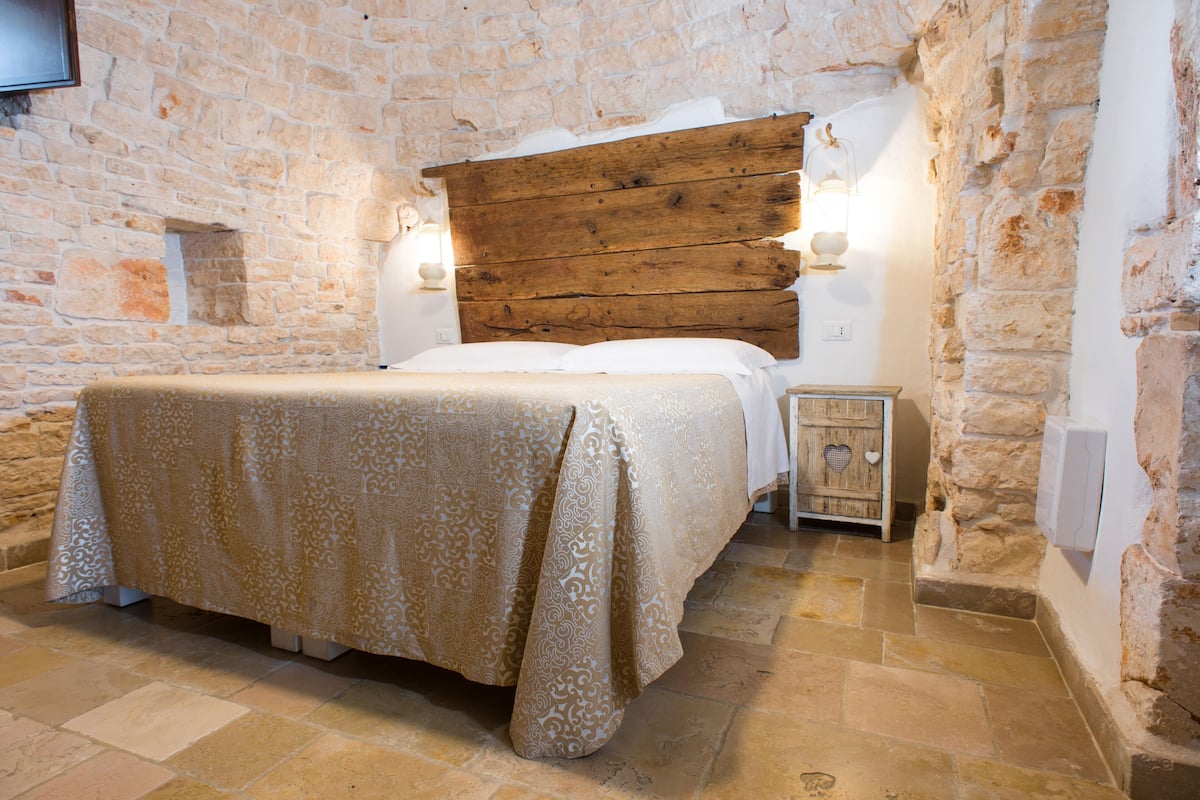 A bedroom with a stone wall, a wooden headboard, a bed with a patterned bedspread, and a nightstand with a lamp.