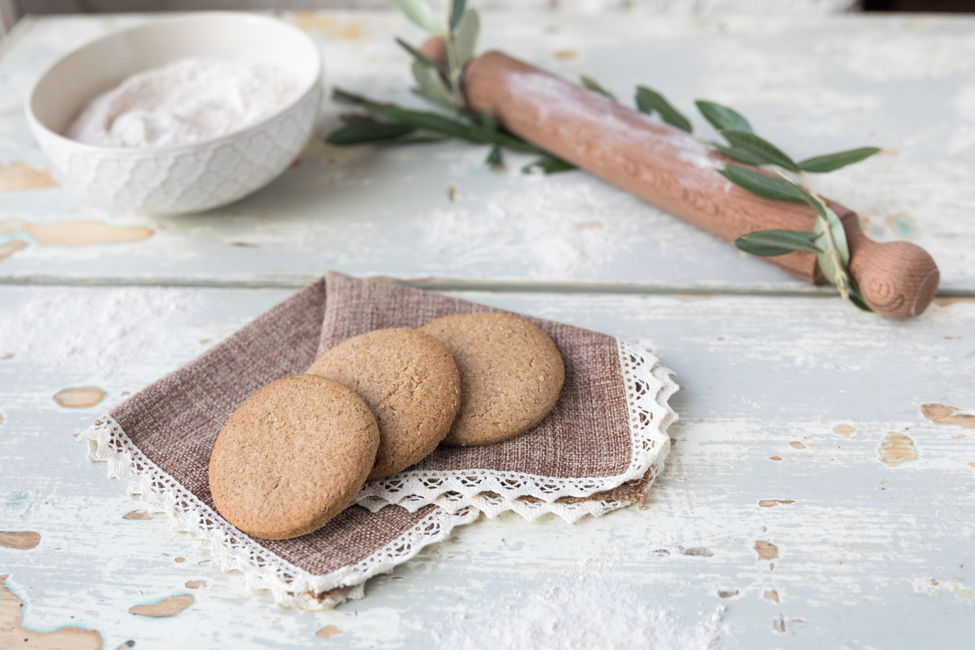 Un décor rustique avec une table en bois sur laquelle sont disposés des biscuits sur un chemin de table en dentelle, un rouleau à pâtisserie et un bol de farine.