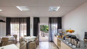 A dining area with tables and chairs, a kitchen counter with coffee makers and fruit, and a view of the outdoors. - Aparthotel Pharia (Hvar)