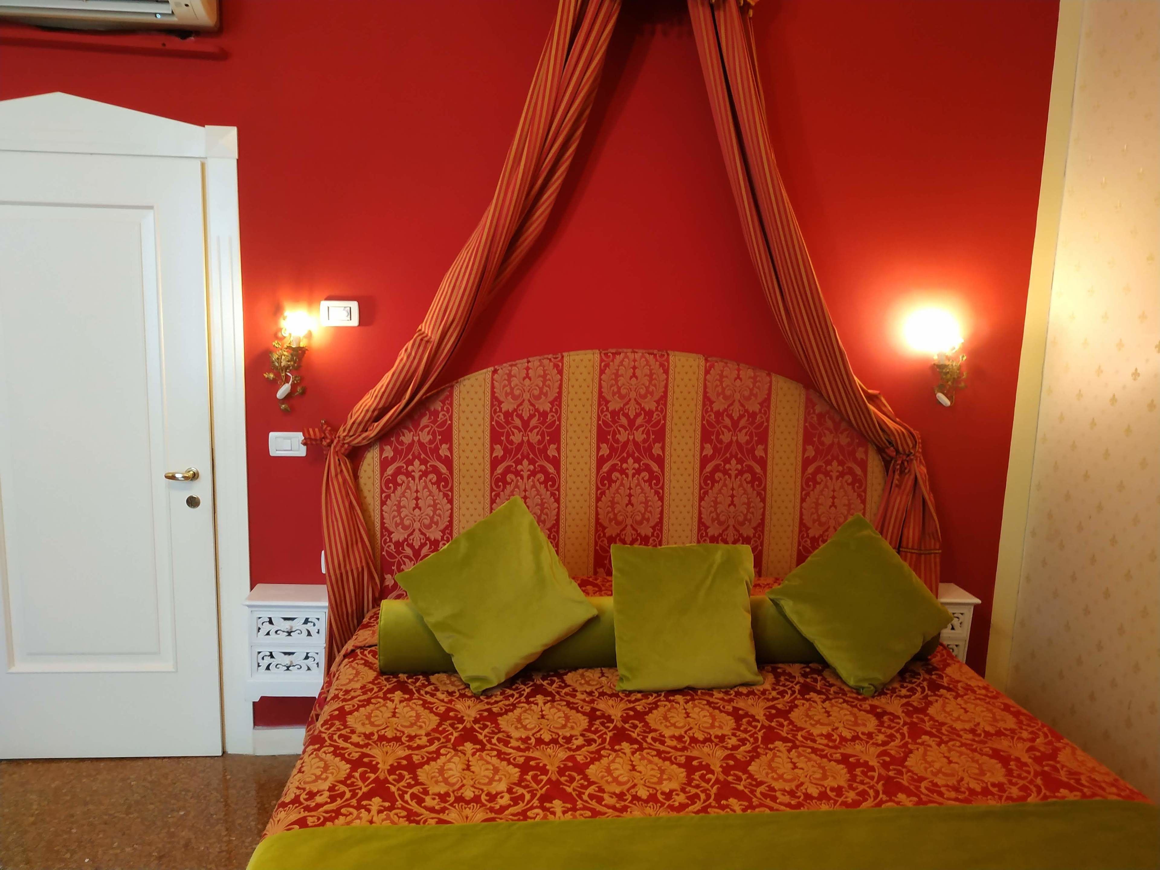 Een kamer met een bed, voorzien van een rood- en goudkleurig beddegoed, groene kussens en een baldakijn. Er zijn wandlampen en een witte deur.