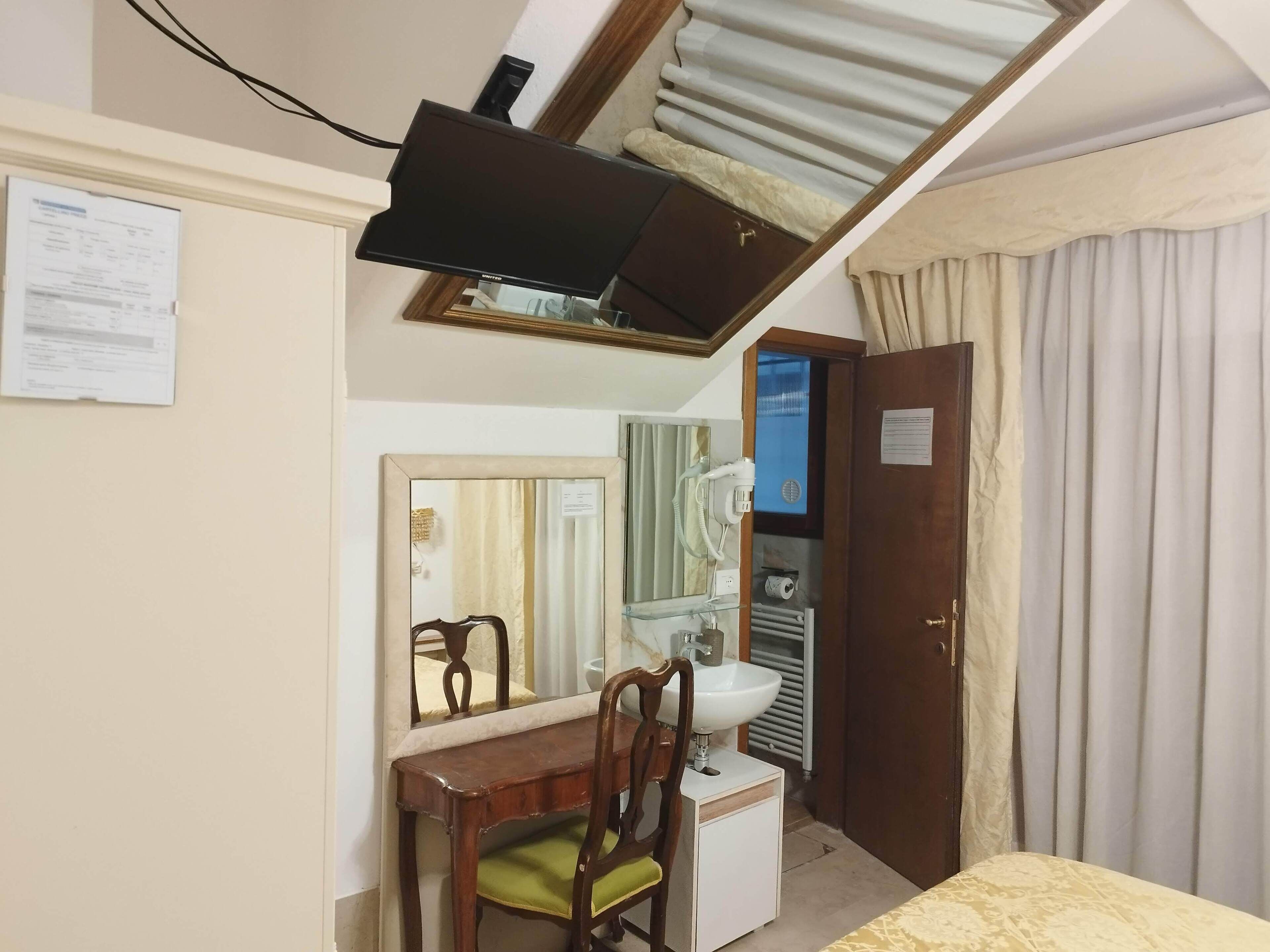 Ein Schlafzimmer mit einem Bett, einem Schreibtisch mit Stuhl, einem Spiegel, einem Fernseher, einer Tür und einem Fenster mit Vorhängen.