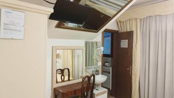 Ein Schlafzimmer mit einem Bett, einem Schreibtisch mit Stuhl, einem Spiegel, einem Fernseher, einer Tür und einem Fenster mit Vorhängen.