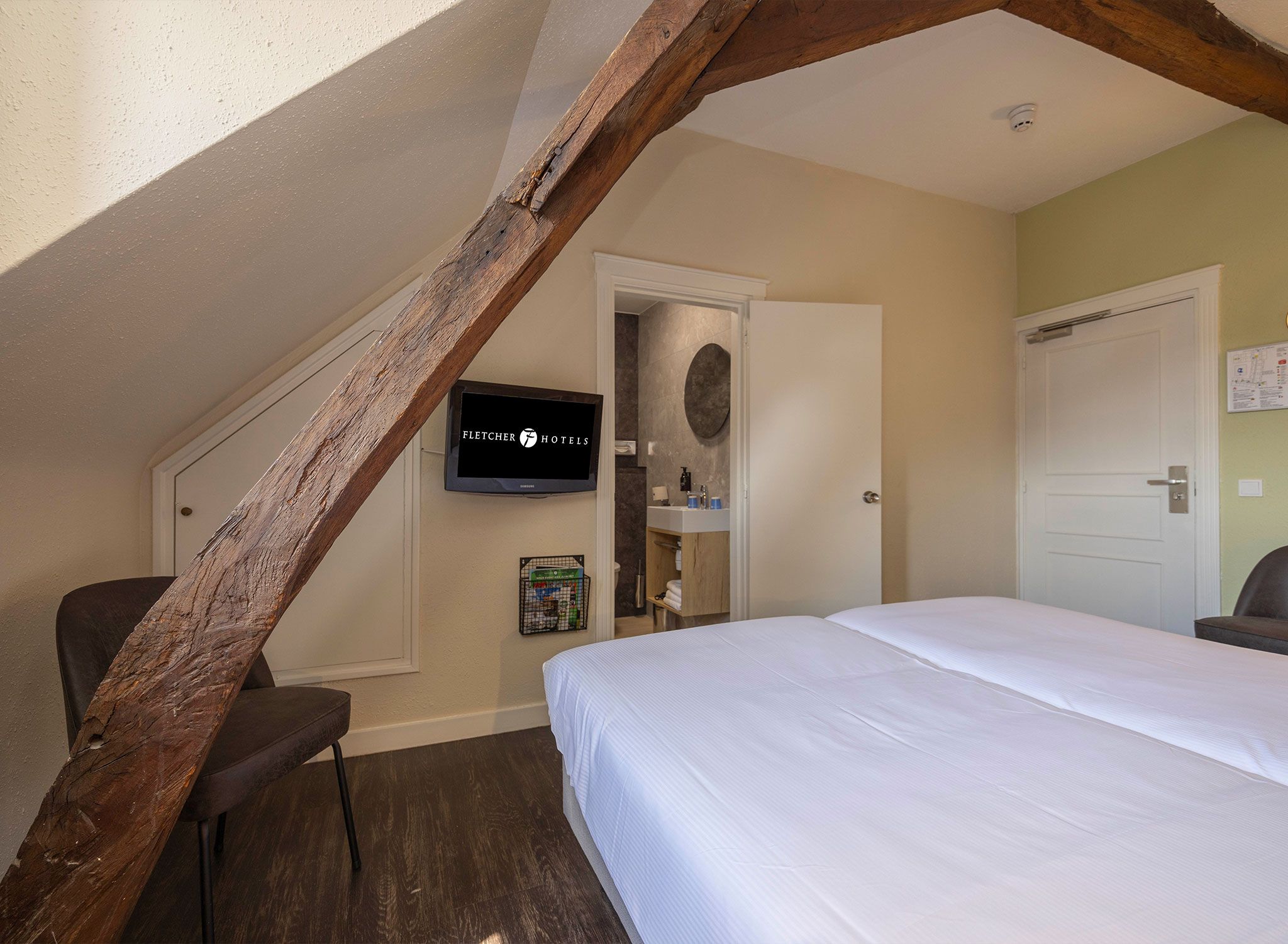 A hotel room with a wooden ceiling beam, a bed, a television, and a bathroom visible through an open door.