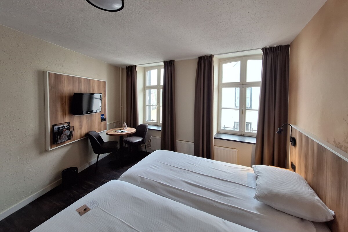 Une chambre d’hôtel avec deux lits, un bureau avec une chaise, une télévision et une fenêtre avec des rideaux.