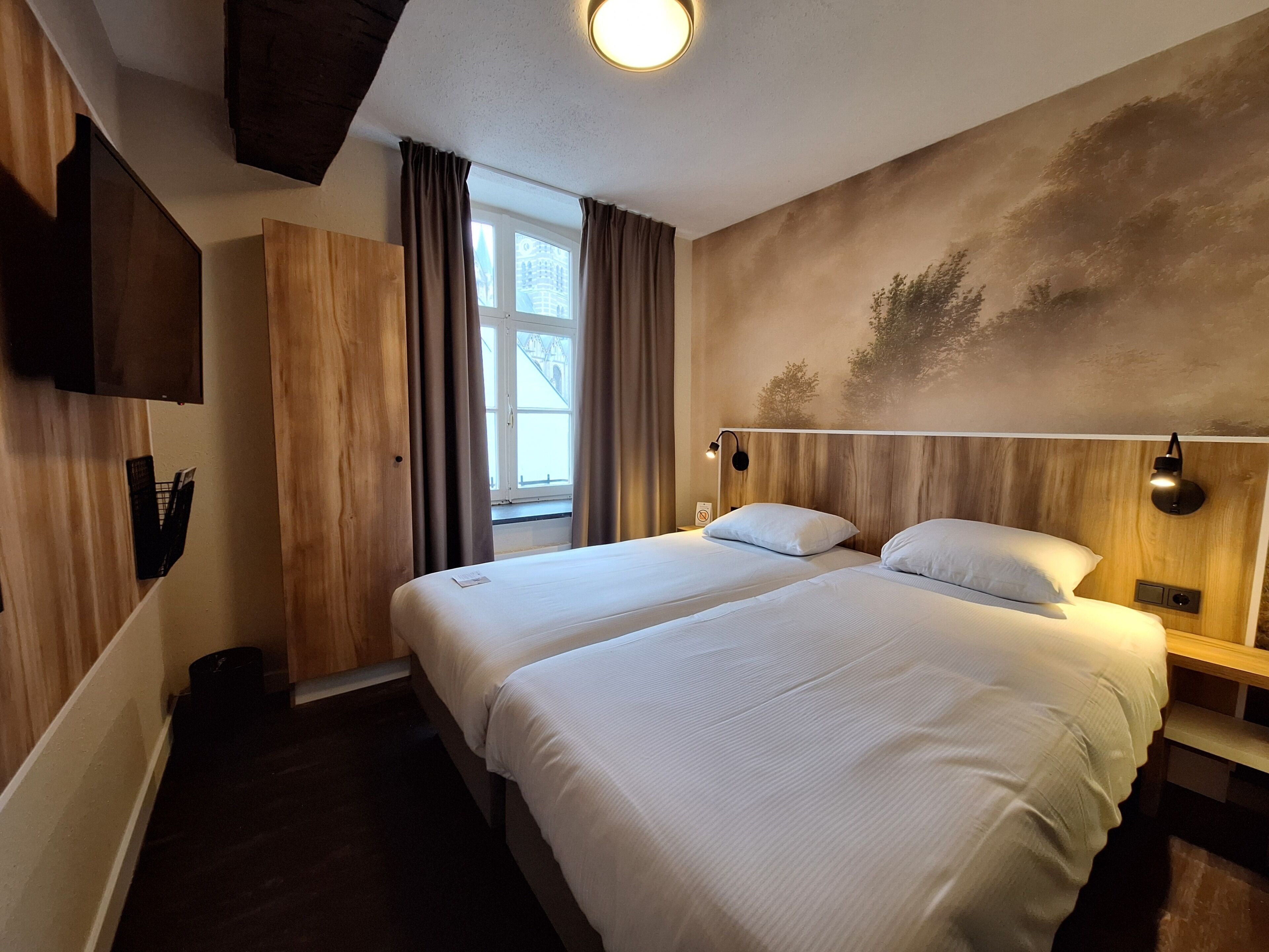 Een hotelkamer met een tweepersoonsbed, een houten hoofdbord, een wandtelevisie en een raam met gordijnen.