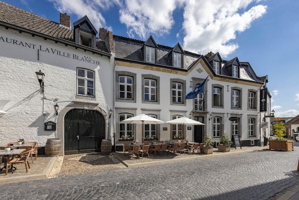 A traditional European building with a sign reading "Restaurant Laville Blanche." It features outdoor seating with tables and chairs, a cobblestone street, and a clear blue sky.