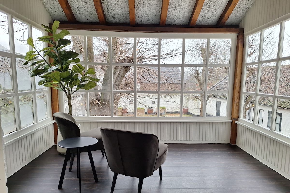Een kamer met een groot raam, een plant in pot, twee stoelen en een tafel.