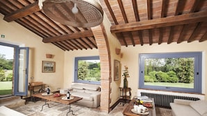 A cozy living room with stone flooring, a wooden ceiling, and large windows offering a view of greenery. - Podere Monti (Montepulciano)