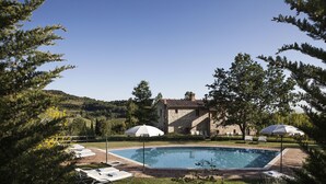 A swimming pool with lounge chairs and umbrellas, surrounded by trees and a stone building in the background. - Podere Monti (Montepulciano)