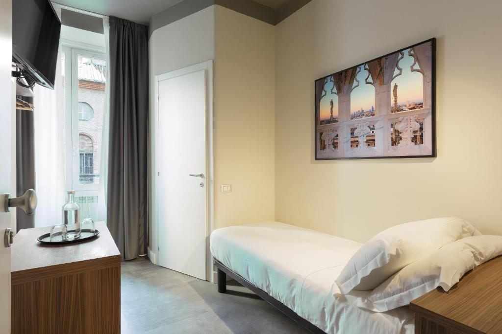 A hotel room with a bed, a bedside table with a tray, a painting on the wall, and a door leading to a balcony.