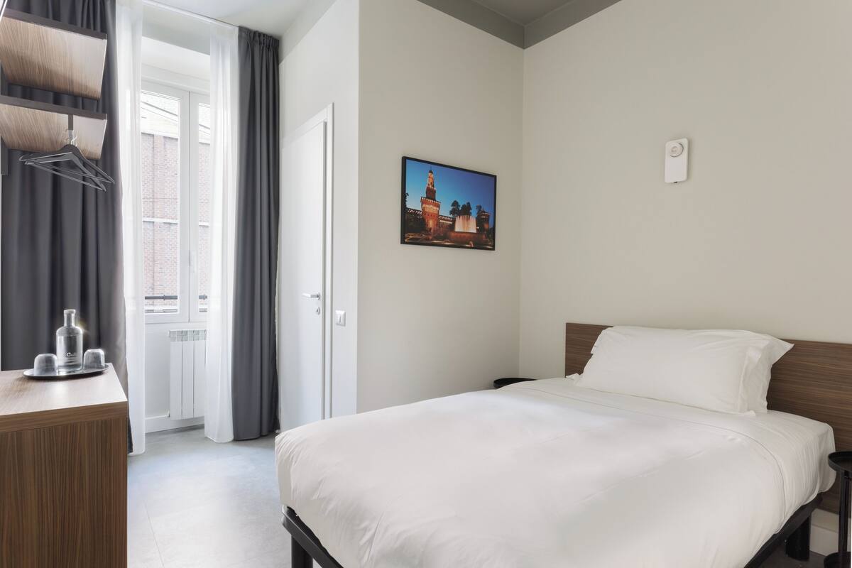 A hotel room with a bed, a desk with a bottle and glasses, a picture on the wall, and a window with curtains.
