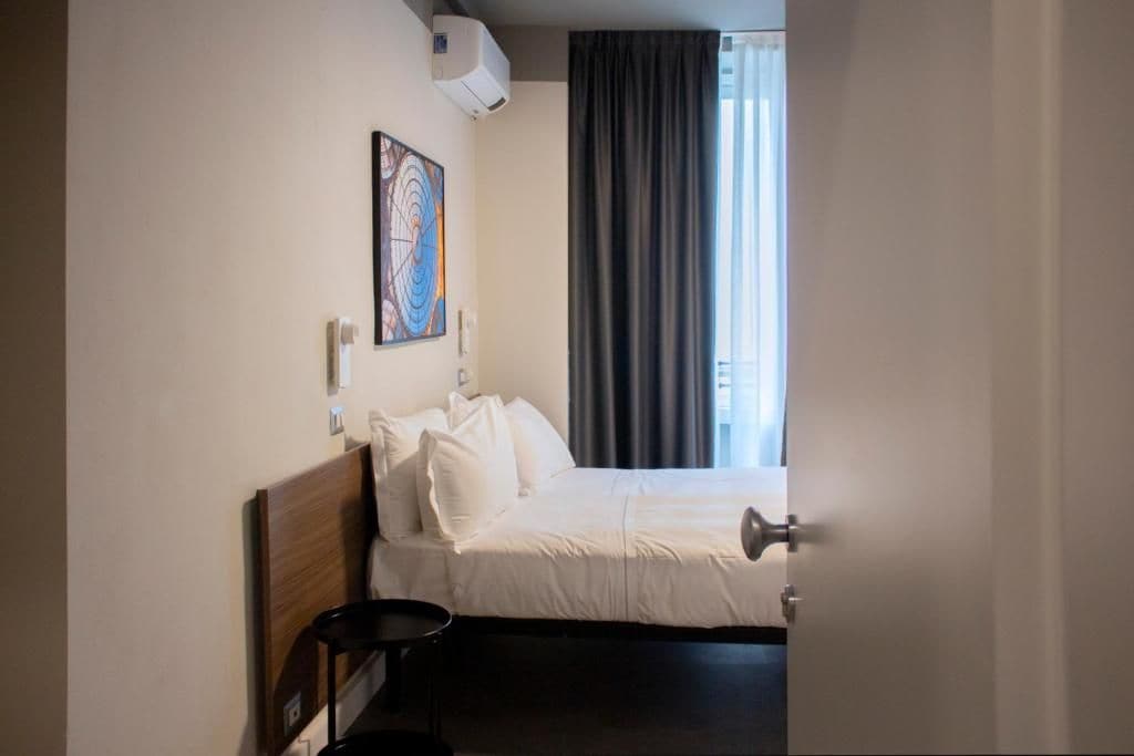 A hotel room with a bed, a bedside table, a wall-mounted air conditioner, a painting, and a window with curtains.