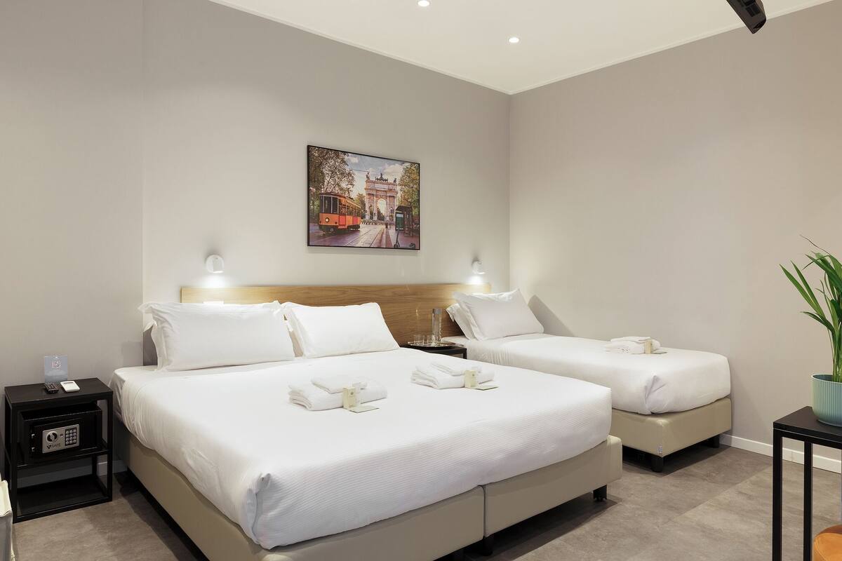 A hotel room with two beds, a painting on the wall, a bedside table with a lamp, and a potted plant.
