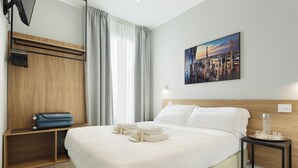 Camera d'albergo con un letto grande, una testiera in legno, un quadro che raffigura un panorama cittadino, un comodino con un bicchiere e una valigia su uno scaffale.