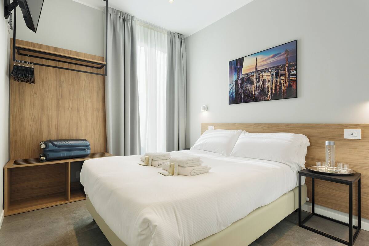 A hotel room with a large bed, a wooden headboard, a painting of a cityscape, a side table with a glass, and a suitcase on a shelf.