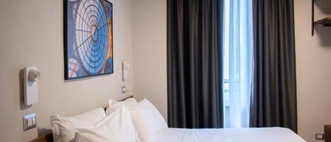 A hotel room with a bed, a wall-mounted air conditioner, a painting, and a window with curtains.
