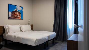 Habitación de hotel con una cama, una mesita de noche, un cuadro de un edificio, una ventana con cortinas y un escritorio con lámpara.