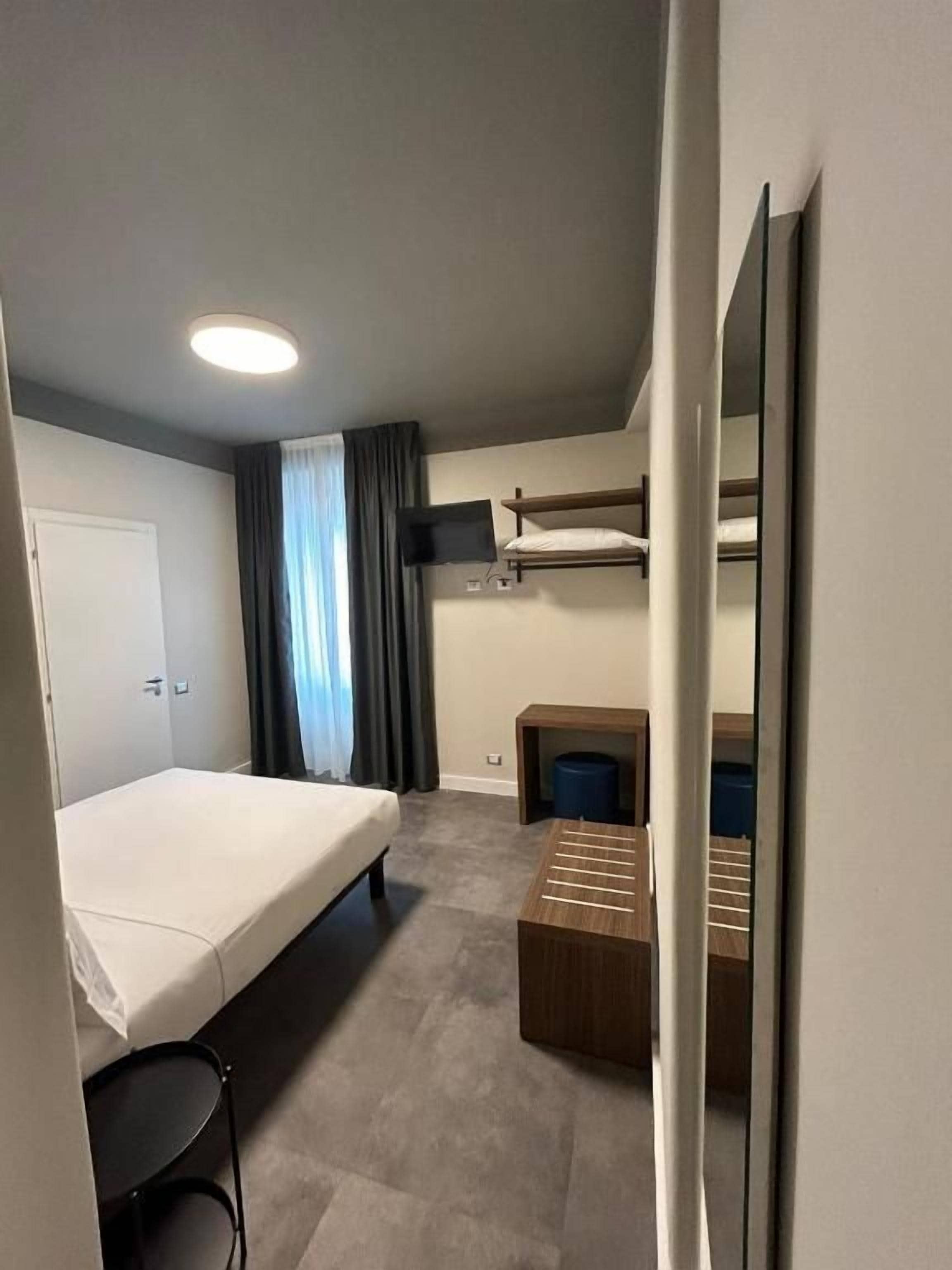 Ein kleines Hotelzimmer mit einem Einzelbett, einem kleinen Schreibtisch, einem Stuhl, einem Spiegel und einem Fenster mit Vorhängen.