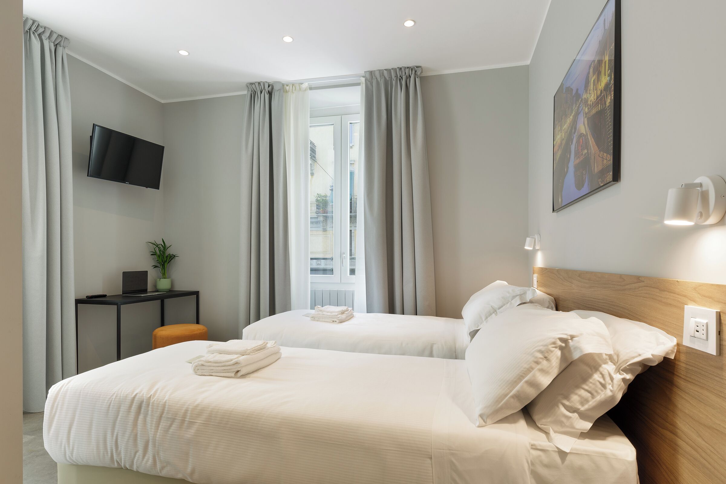 A hotel room with two beds, a flat-screen TV, a desk with a plant, and a window with curtains.