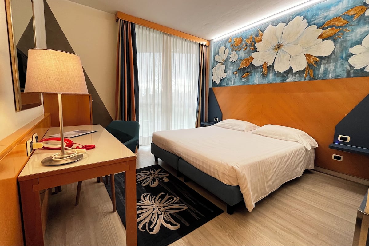 A hotel room with a bed, a desk, a lamp, a telephone, and a painting on the wall.