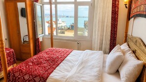 Een slaapkamer met een groot raam, een bed met witte lakens, een houten garderobe en een rood bedovertrek met een patroon.