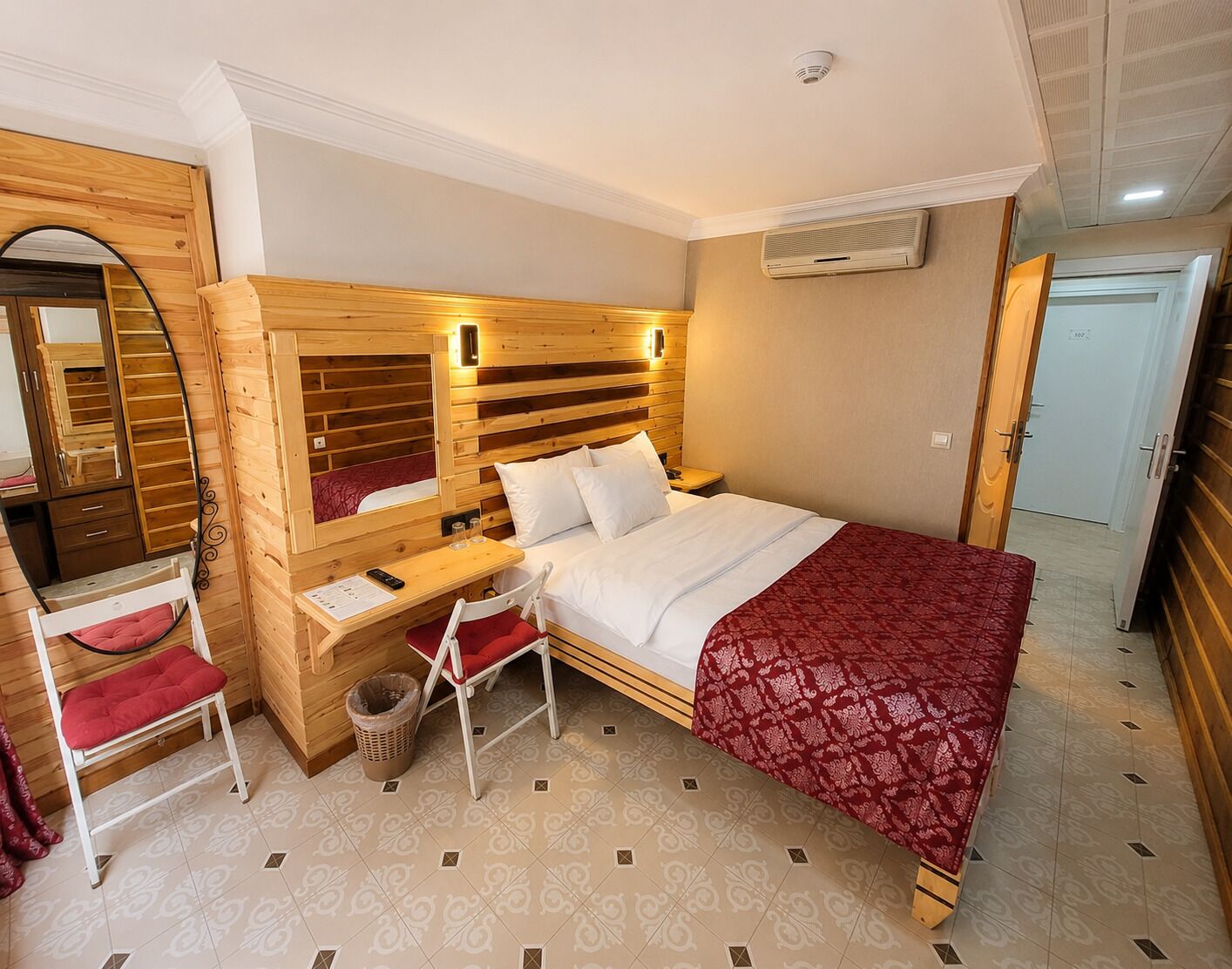 Une chambre à coucher avec une tête de lit en bois, un bureau avec une chaise, un lit recouvert d’une couverture rouge, un miroir et une armoire.