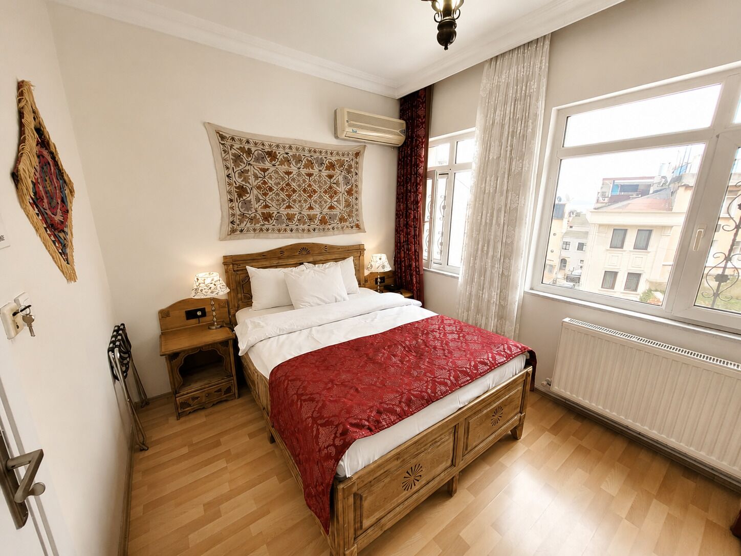 Een slaapkamer met een houten bed, een nachtkastje, wandtapijt en een raam met uitzicht op gebouwen.