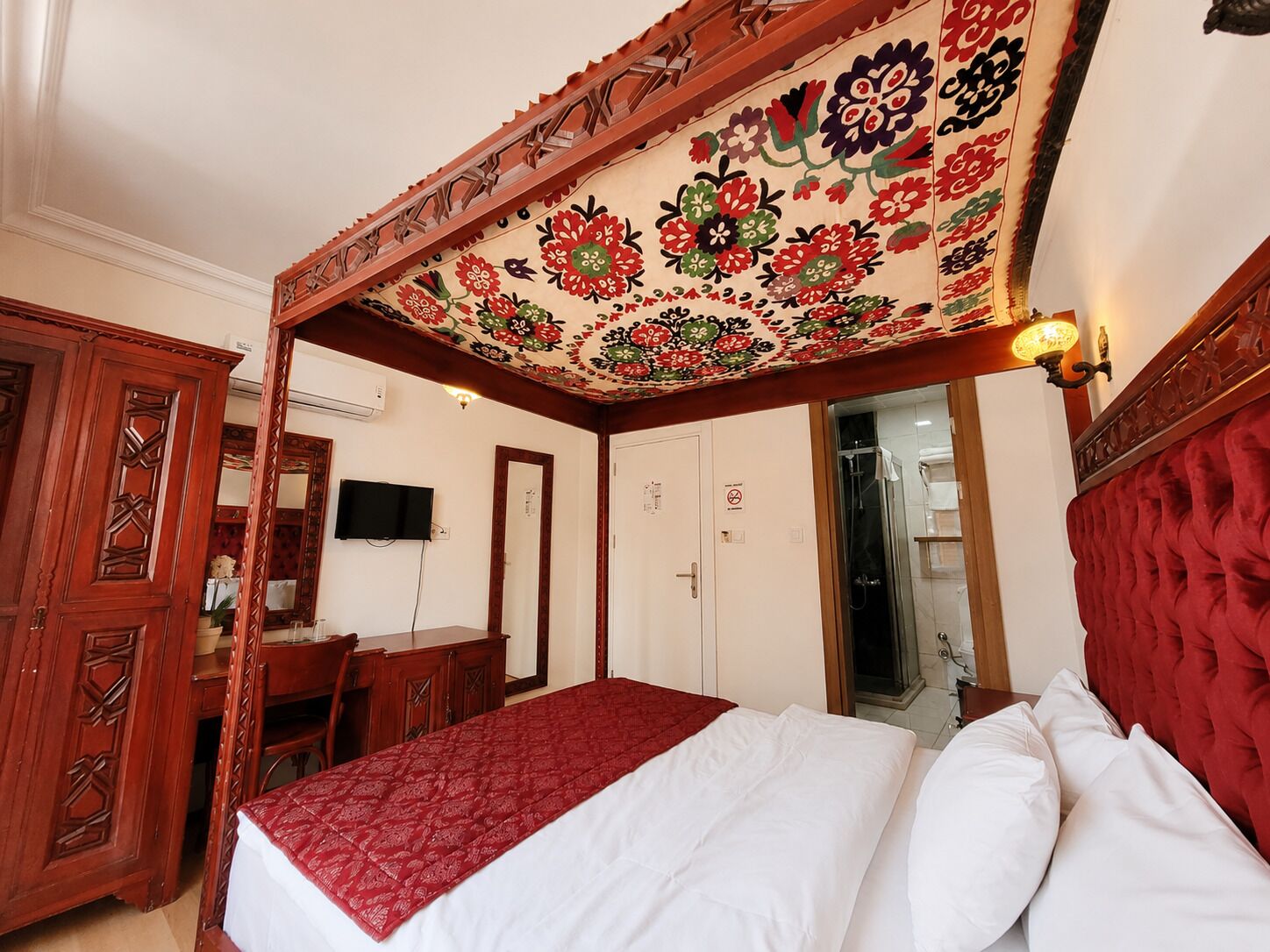 A four-poster bed with a red bedspread, a wooden wardrobe, a desk with a chair, and a television mounted on the wall.