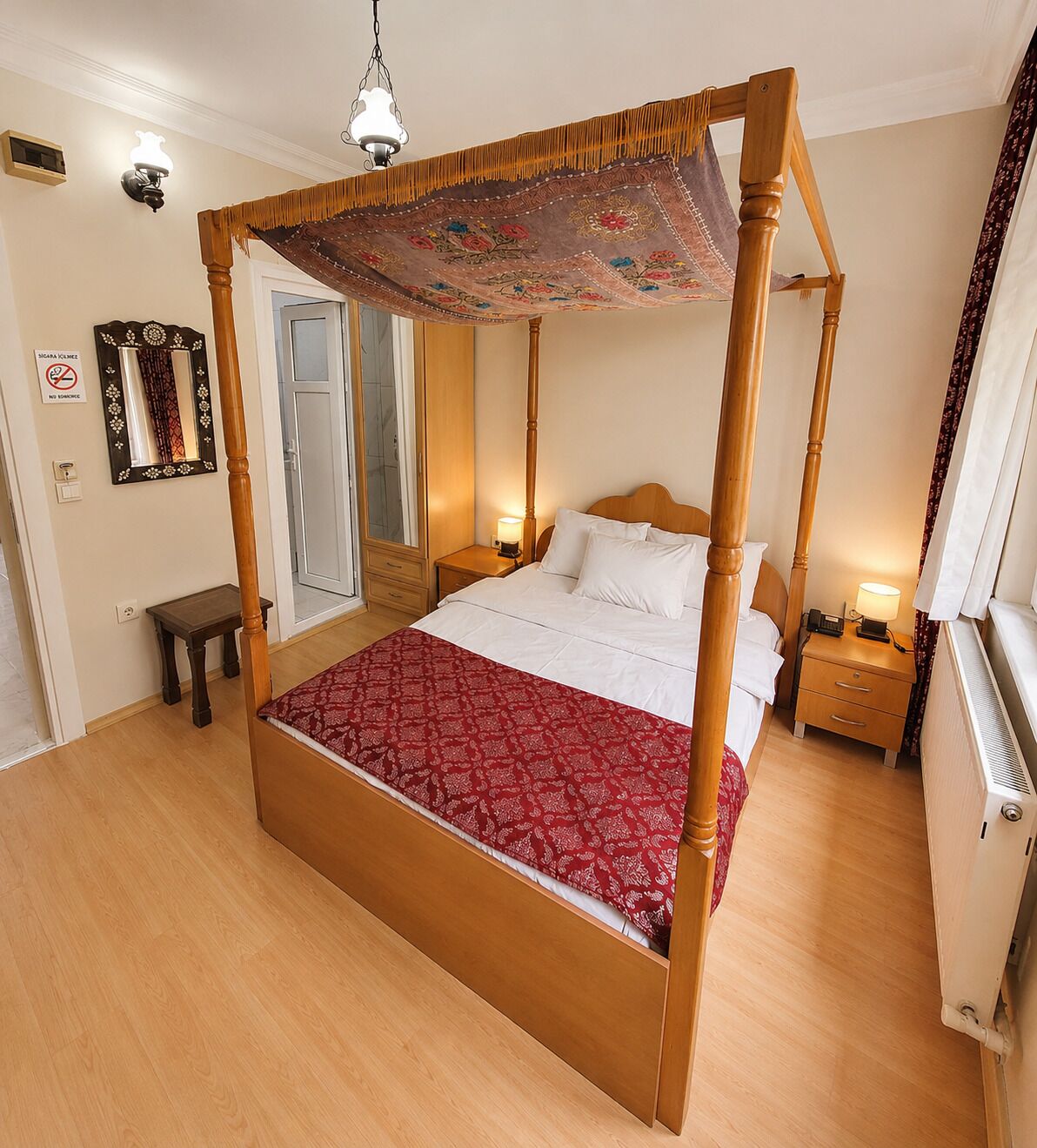 A four-poster bed with a red patterned bedspread, wooden nightstands with lamps, a mirror, and a radiator.