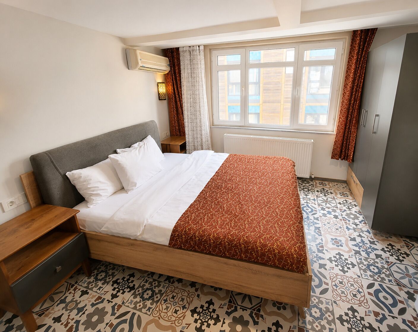 A neatly made bed with a patterned bedspread, a bedside table, a wardrobe, and a window with curtains.