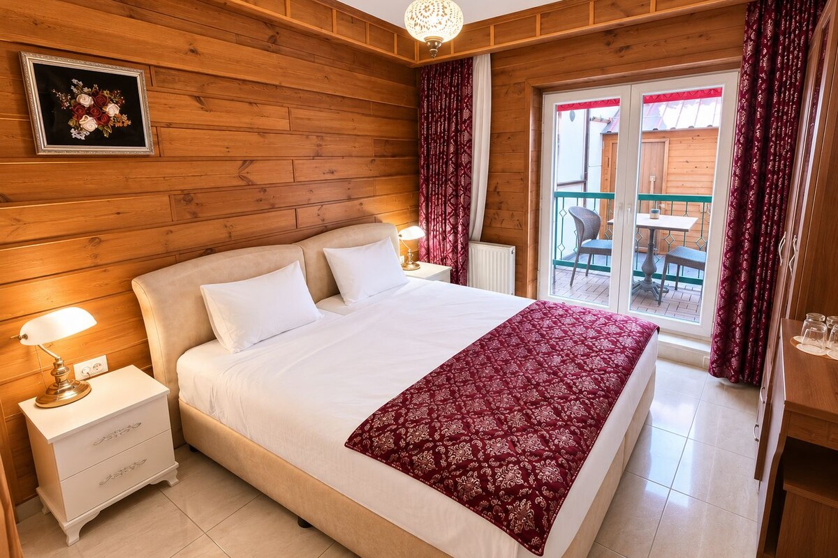 Une chambre d’hôtel avec un mur en bois, un lit avec une literie blanche, un plaid à motifs rouges, une table de chevet avec une lampe et une vue sur le balcon extérieur.
