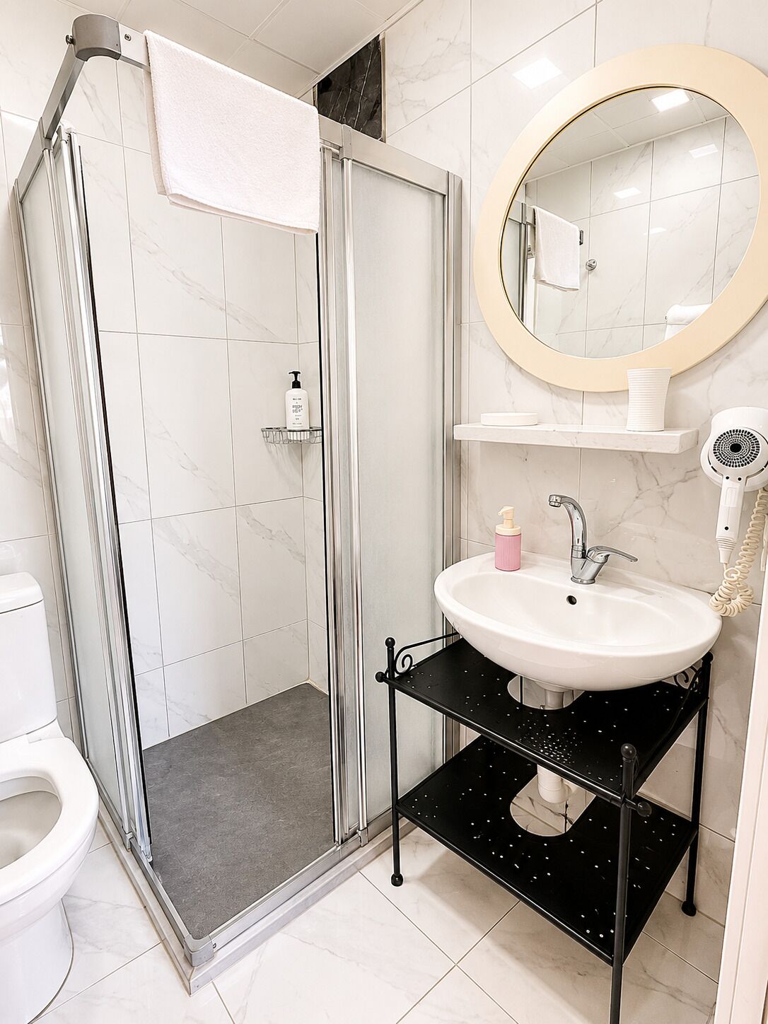 A bathroom with a glass shower enclosure, a white sink with a black shelf, and a round mirror with a beige frame.