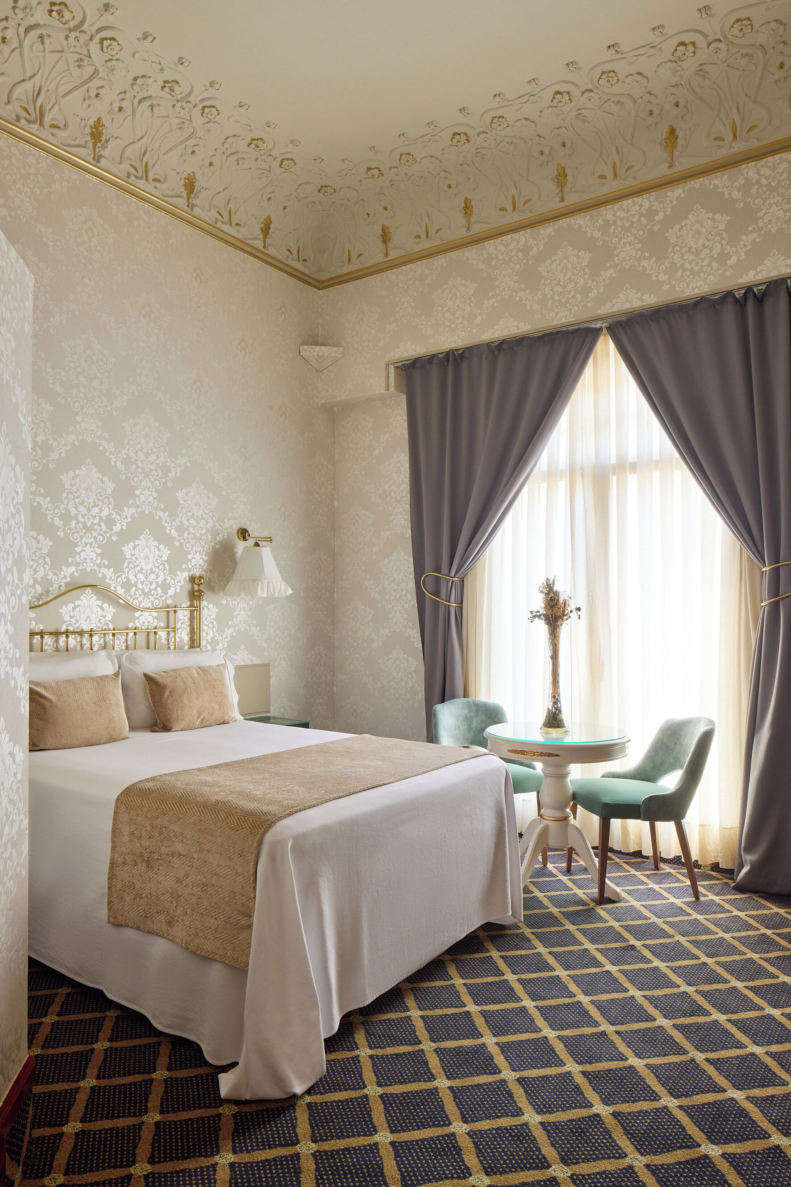 Ein Hotelzimmer mit einem großen Bett, einem kleinen Tisch mit einer Vase, zwei Stühlen und einem gemusterten Boden.