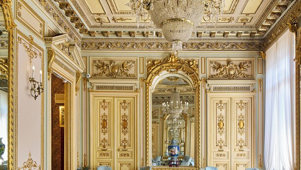 Un comedor elegante con candelabro, techo ornamentado y un gran espejo.