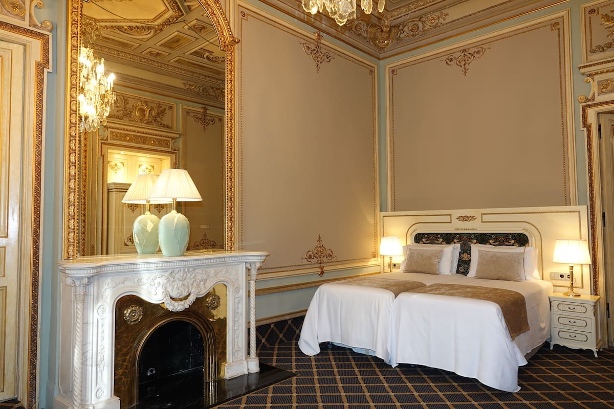 Une chambre d’hôtel luxueuse, dotée d’un grand lit, d’une cheminée, de décorations murales raffinées et d’un lustre.