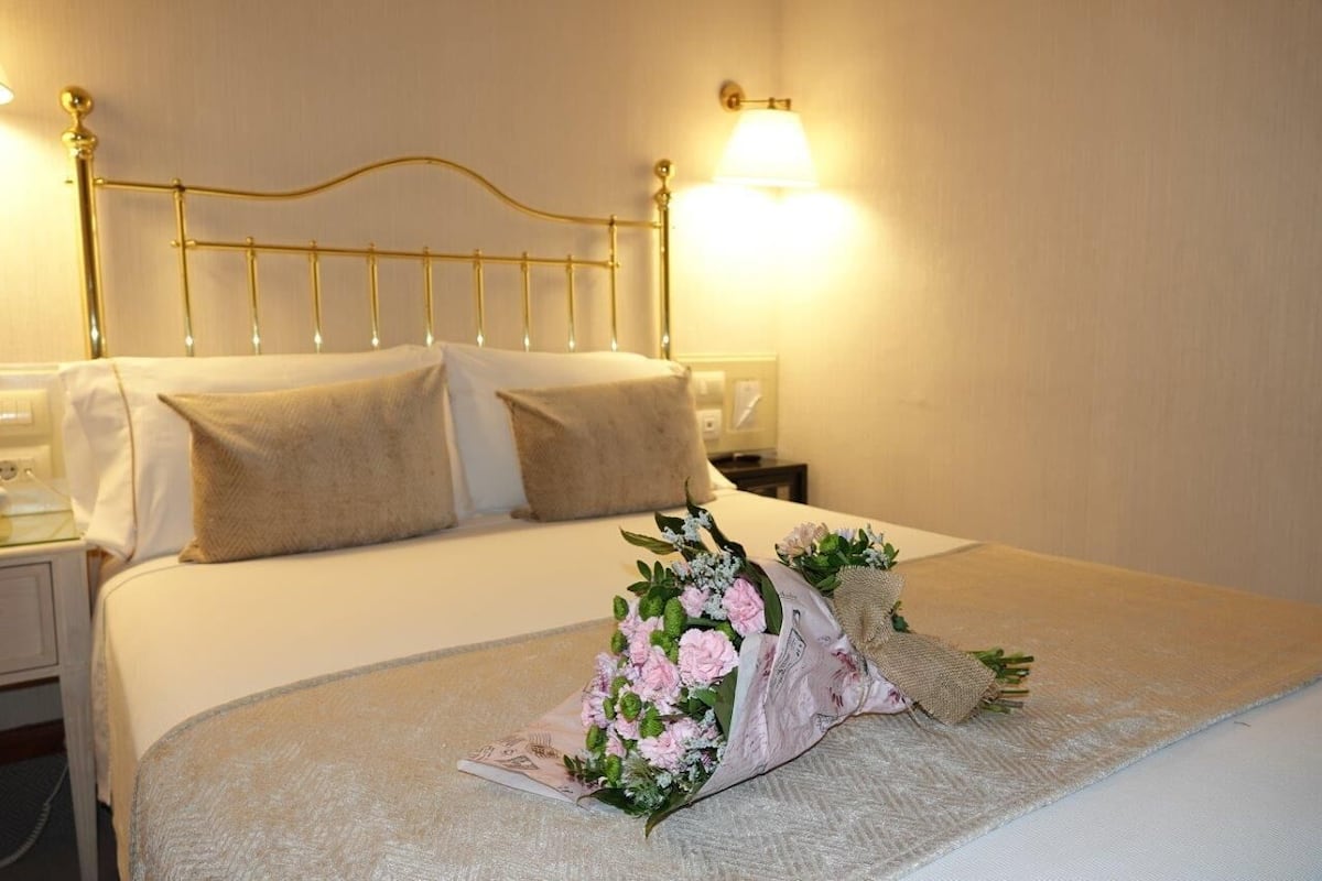 Un lit bien fait, avec une tête de lit, une table de chevet avec une lampe et un bouquet de fleurs posé sur le lit.