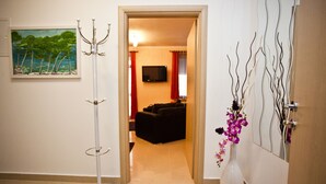 En hall med ett vitt golv, en målning av ett landskap, en vit vas med lila blommor och en svart soffa i bakgrunden.