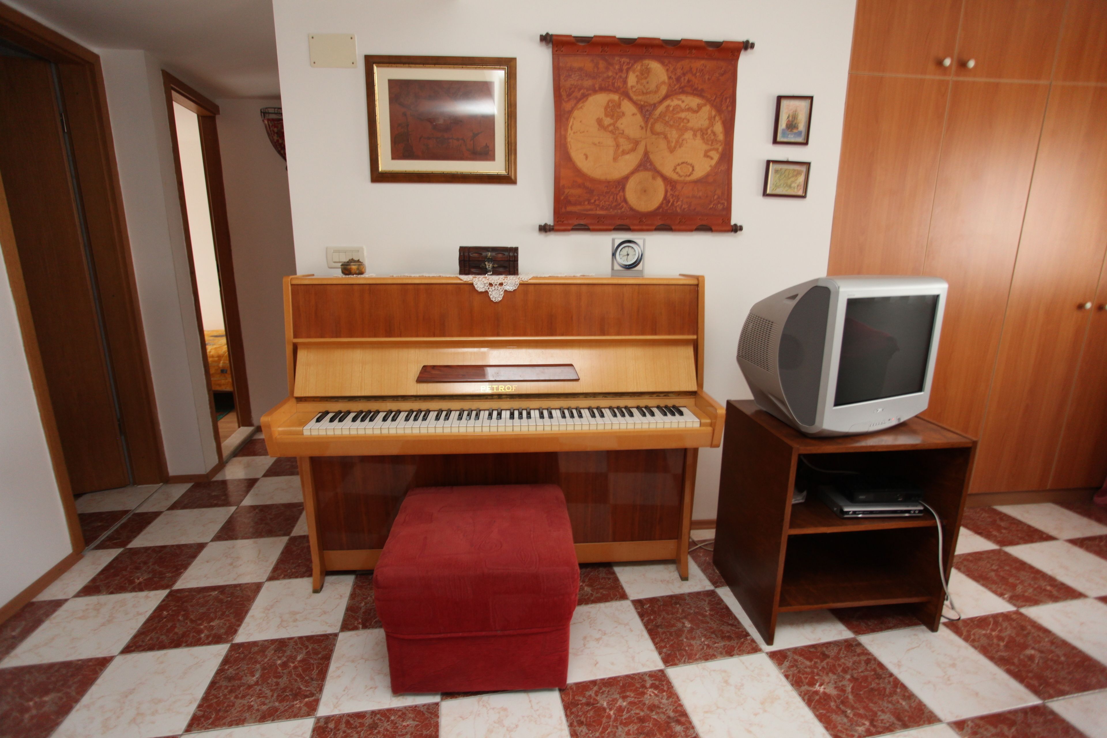 A room with a wooden piano, a red ottoman, a television on a wooden stand, and a patterned tapestry on the wall.