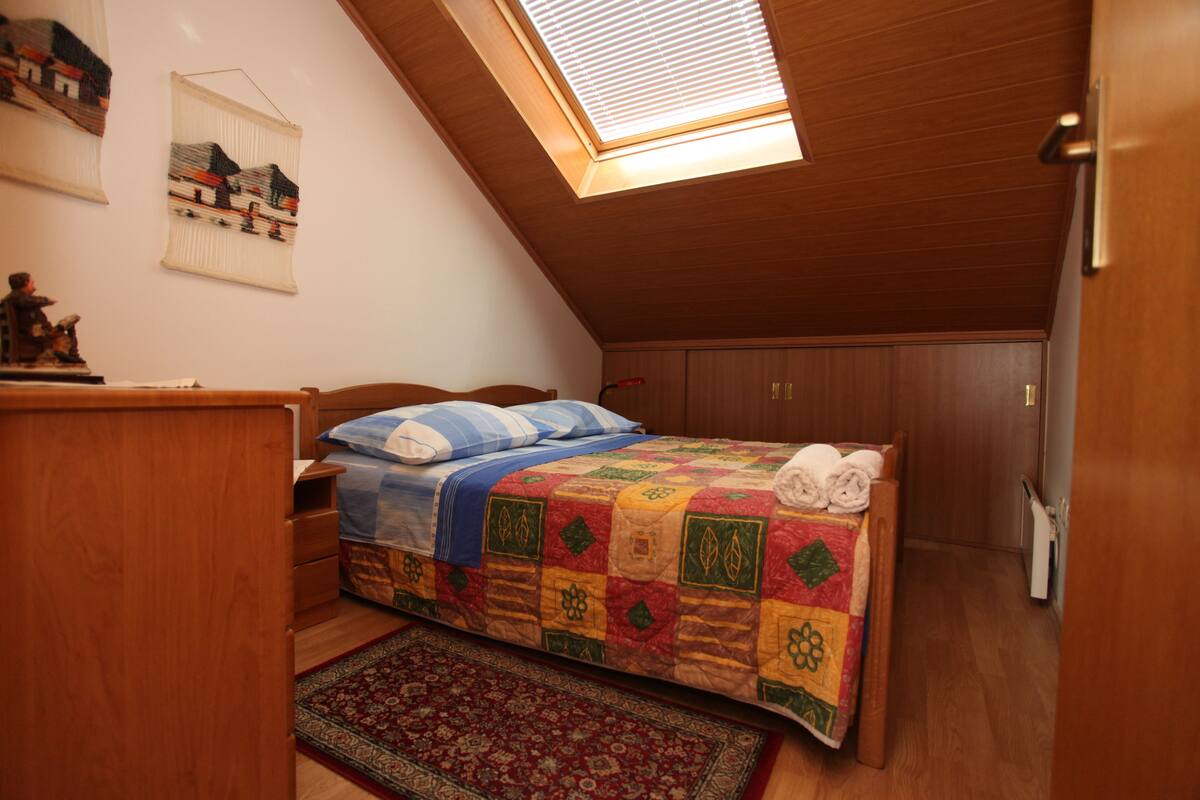A bedroom with a wooden ceiling, a skylight, a bed with a colorful quilt, a nightstand, a dresser, and a small statue.