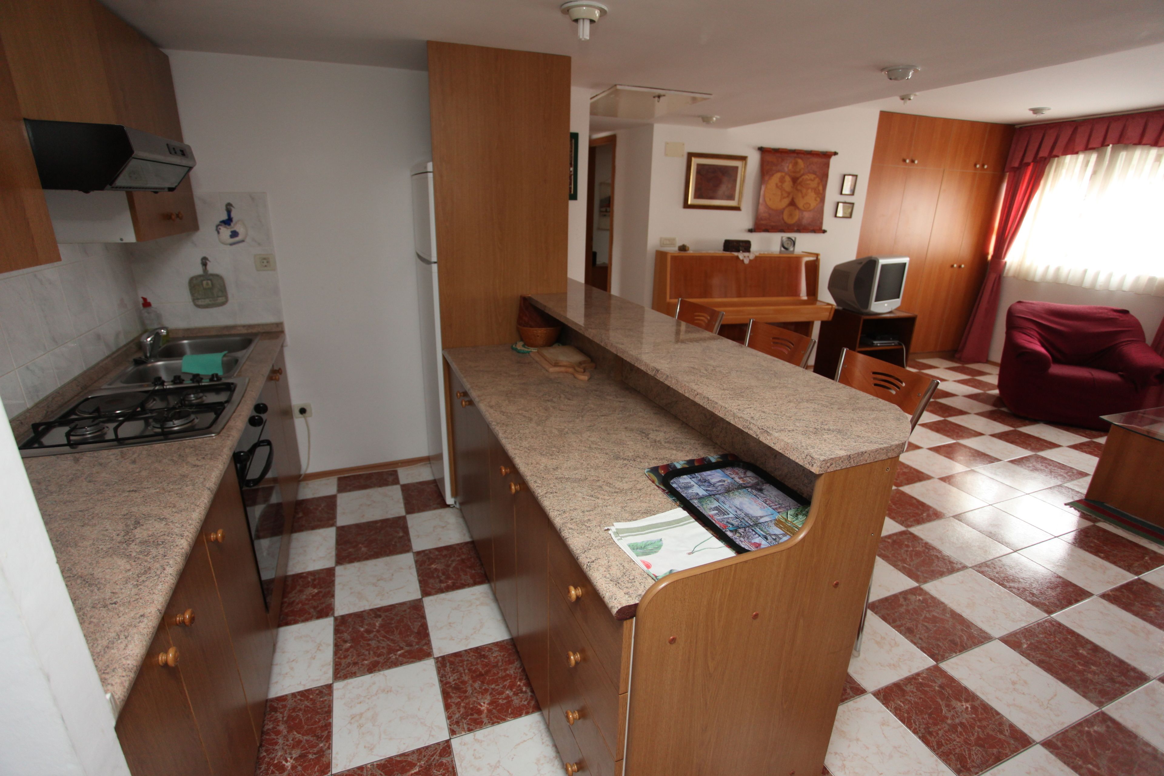 Una cocina compacta con estufa a gas, un lavamanos y piso a cuadros. Hay una zona de comedor con una mesa y sillas, y un sillón rojo.