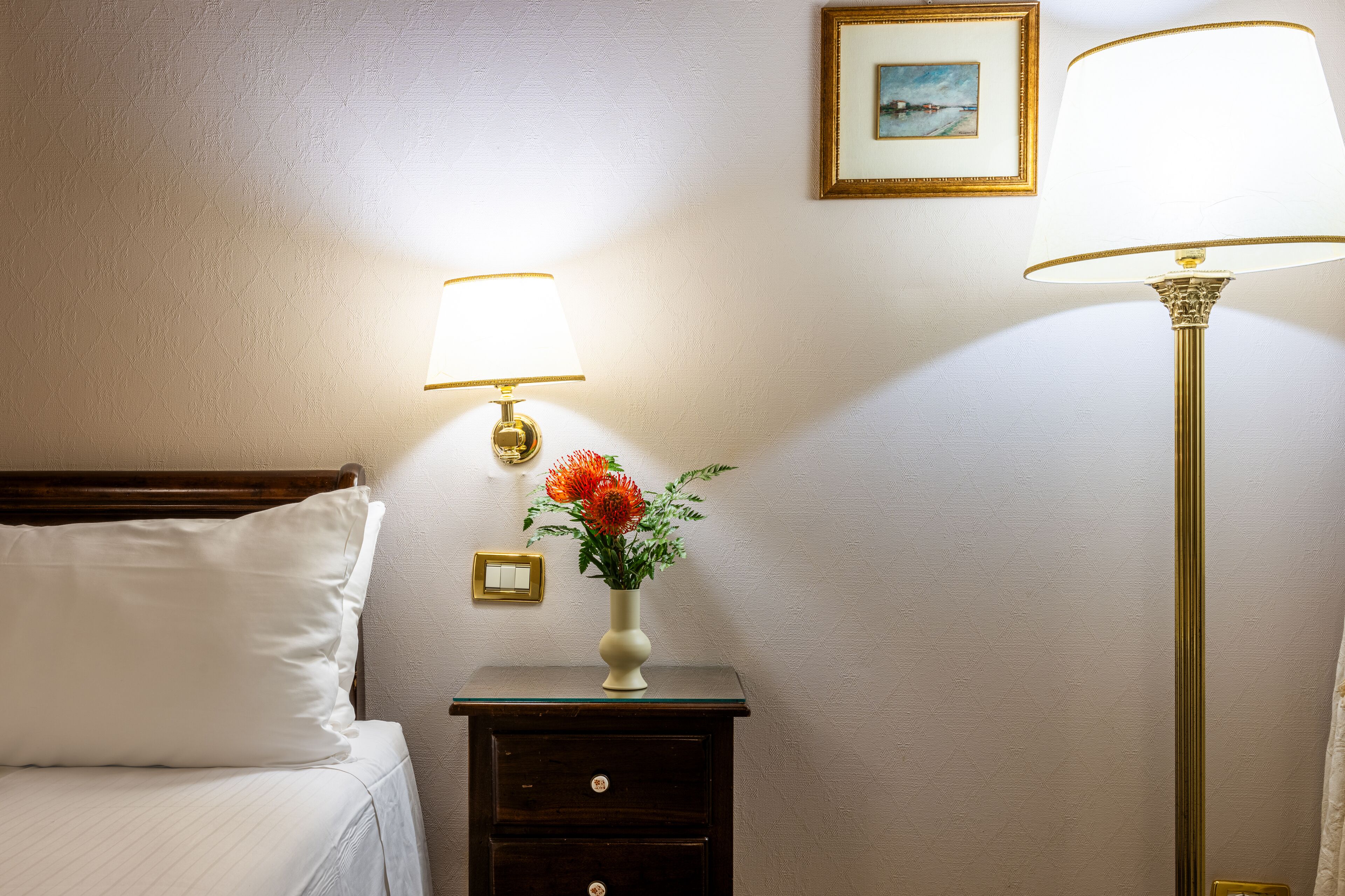A neatly made bed with white linens, a small bedside table with a lamp, a vase with flowers, and a framed picture on the wall.