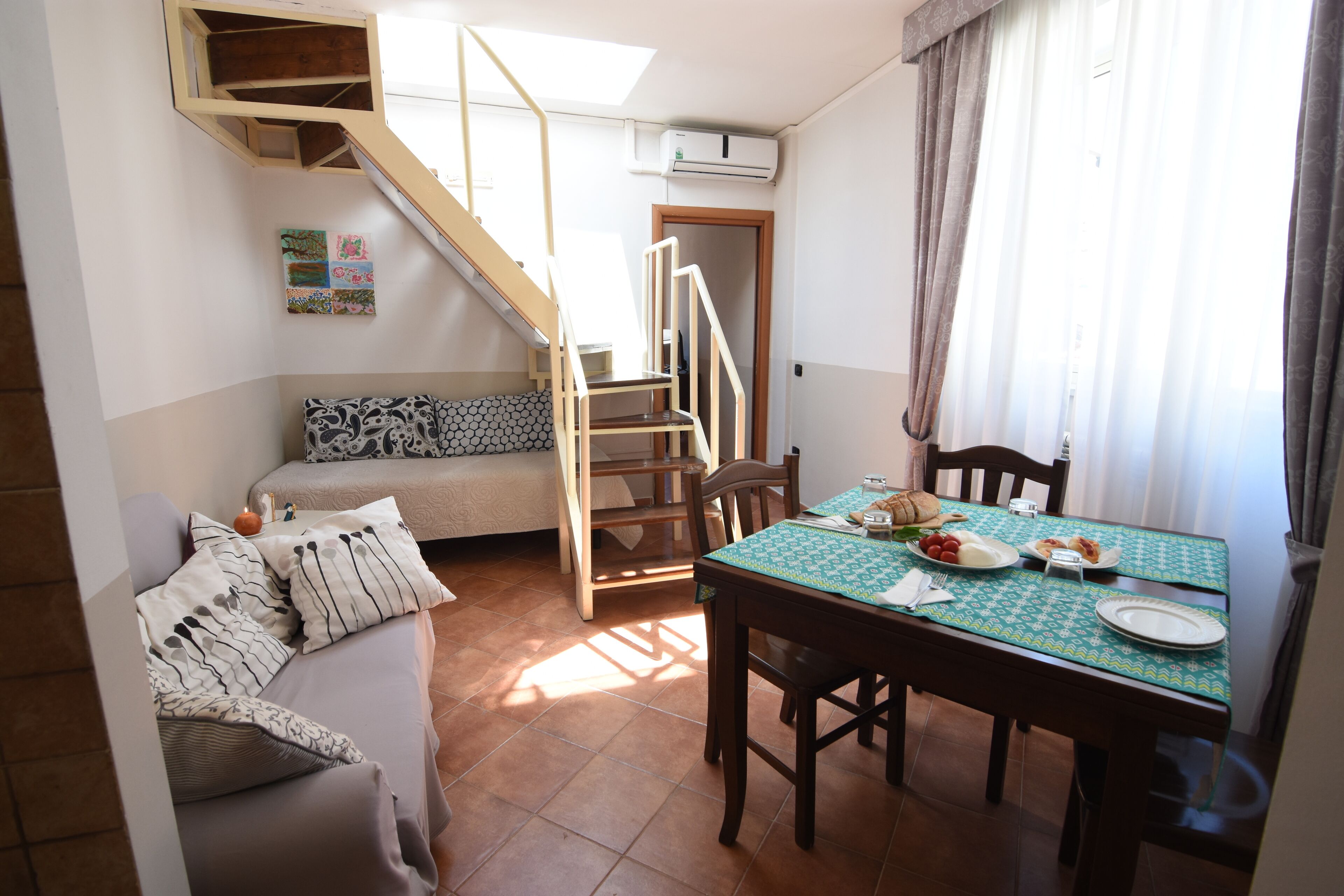 Une pièce chaleureuse avec une table à manger dressée pour deux personnes, un canapé agrémenté de coussins à motifs, un escalier et une petite fenêtre avec des rideaux.